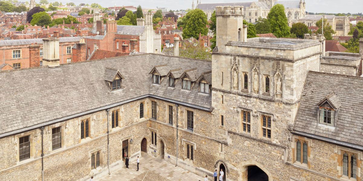 Aerial view of a historic stone building with a slate roof, multiple windows, and an arched entrance, surrounding a central courtyard, with additional buildings and trees in the background.